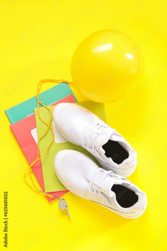 Sneakers with whistle, ball and notebooks on yellow background