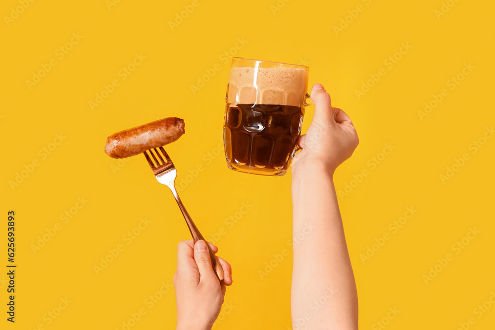 Female hands with mug of cold beer and sausage on yellow background