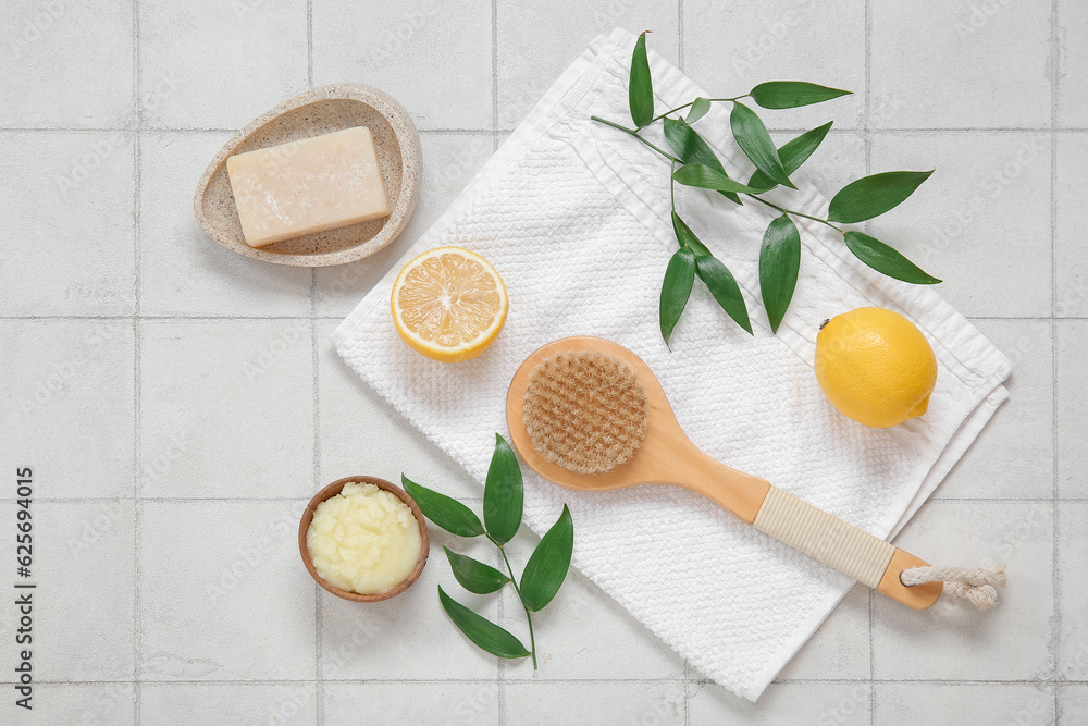 Bowl of lemon body scrub with massage brush, towel and soap on white tile background