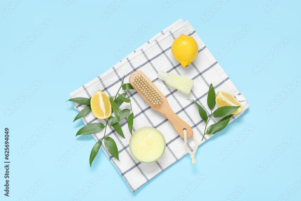 Bowl of lemon body scrub with massage brush and cosmetic bottle on blue background