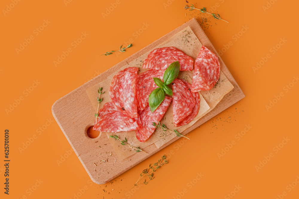 Wooden board with slices of tasty salami on orange background