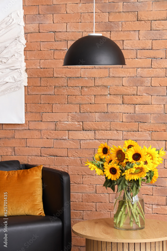 Vase with beautiful sunflowers on coffee table and black armchair near brown brick wall