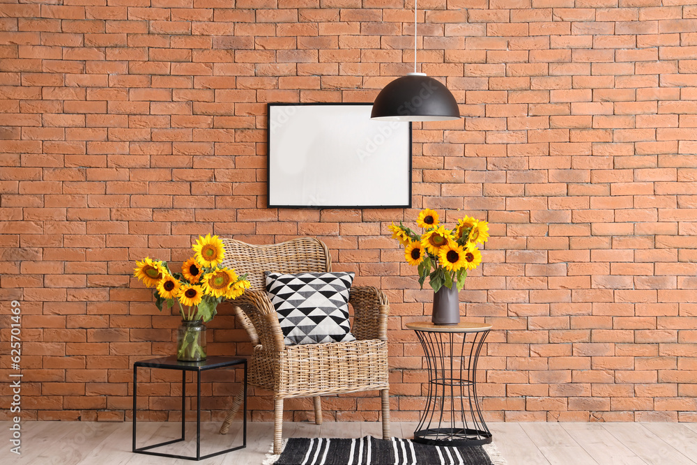 Vases with beautiful sunflowers on coffee tables and wicker armchair near brown brick wall