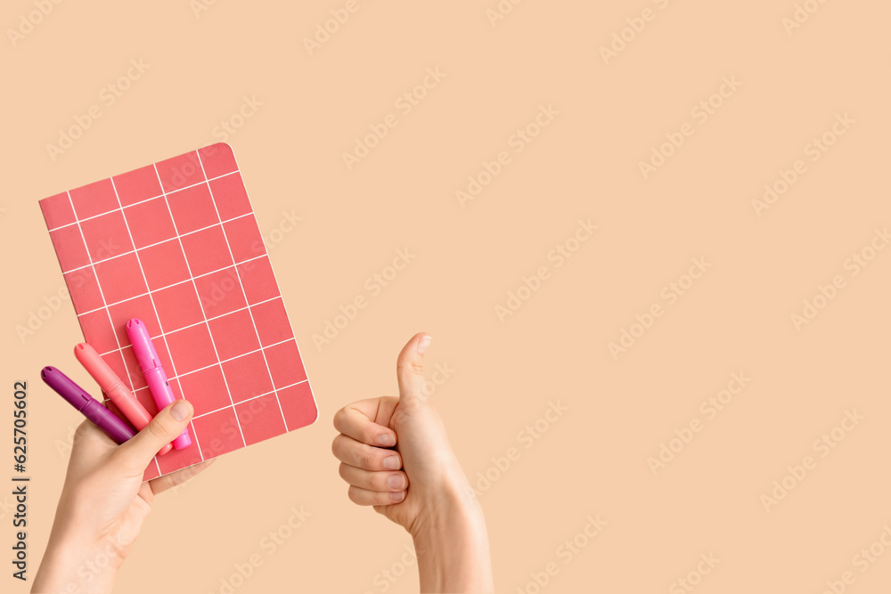 Female hands with notebook and felt tip pens showing thumb up on beige background