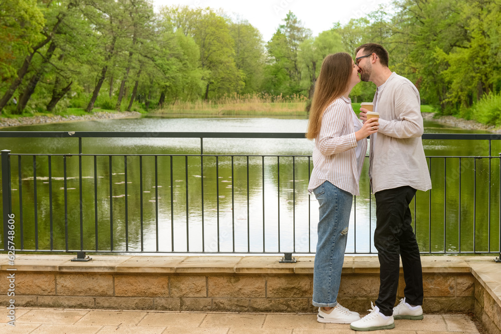 Beautiful happy couple with cups of coffee kissing on bridge in park