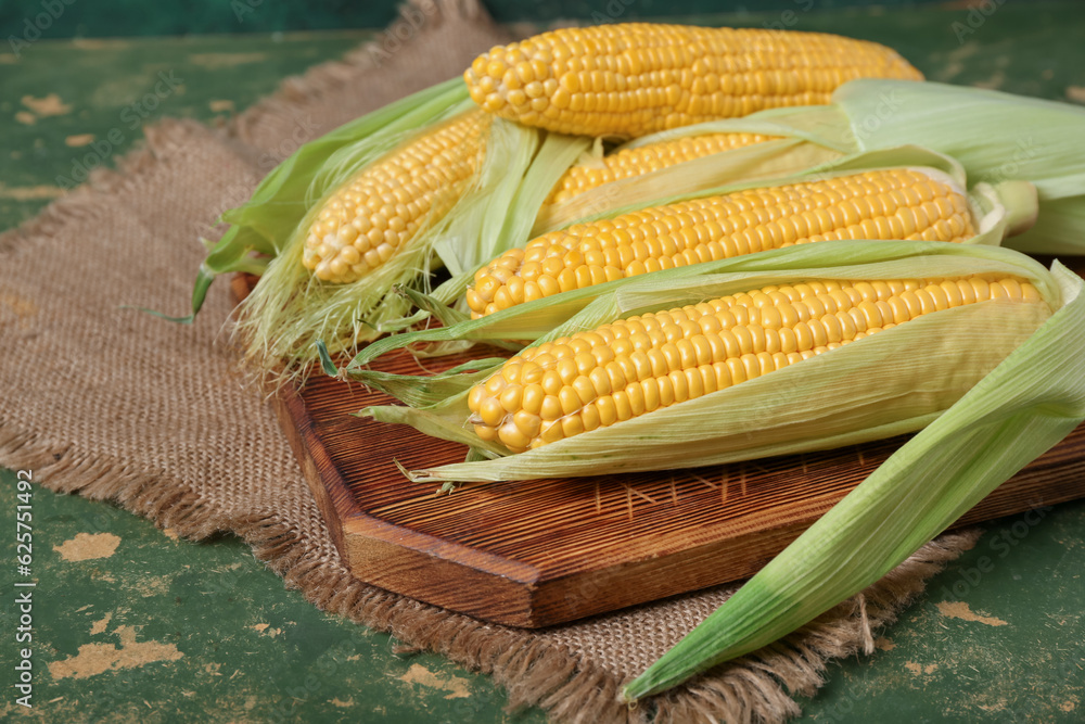 Wooden board with fresh corn cobs on green background