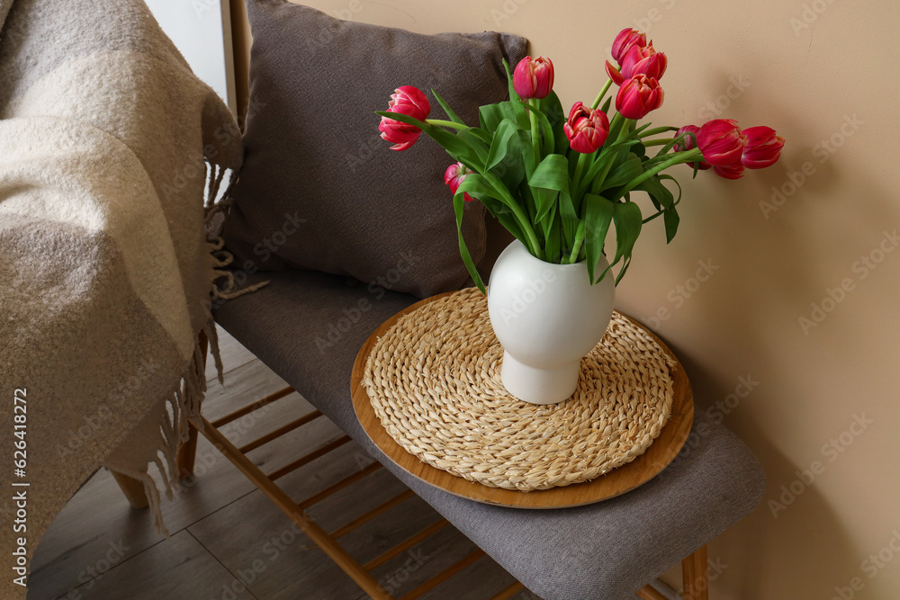 Vase with tulip flowers and cushion on grey bench near beige wall, closeup