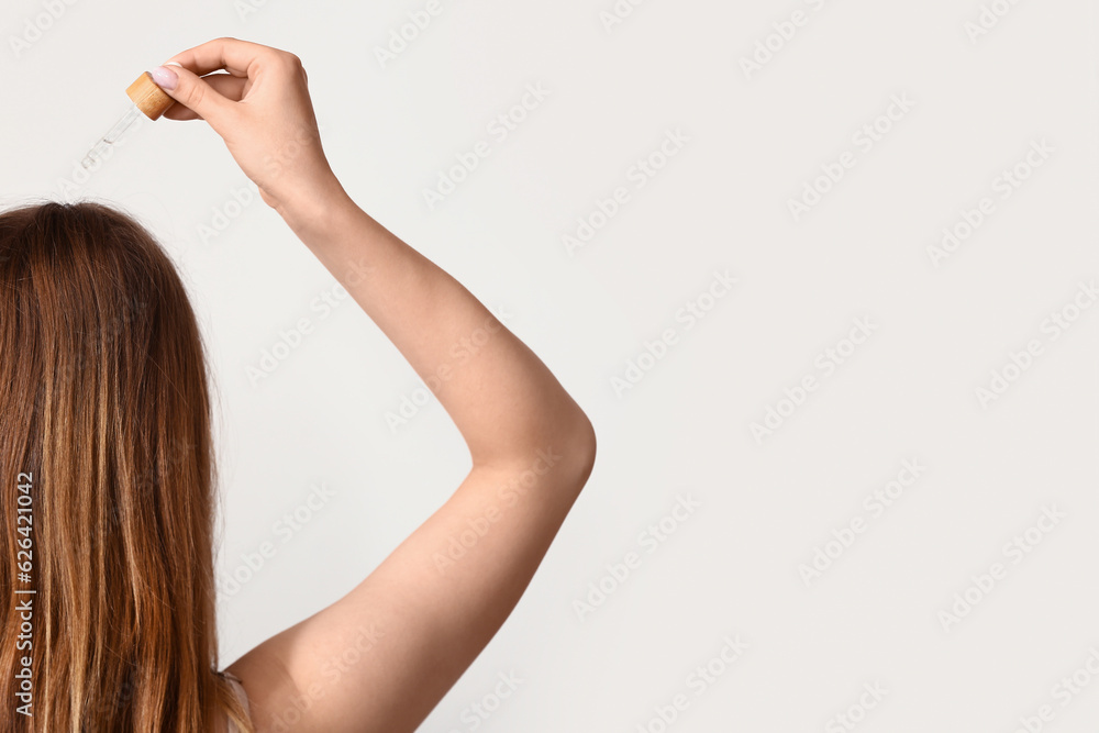 Young woman using cosmetic oil for hair treatment on light background, back view