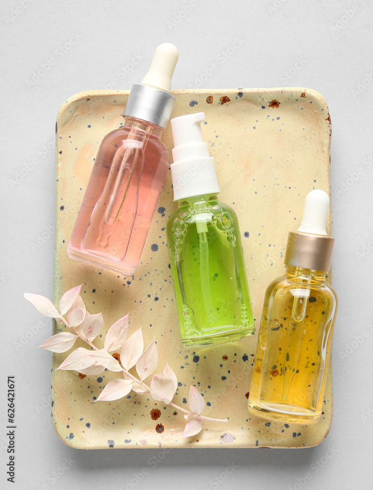 Plate with bottles of cosmetic products and dried plant on light background