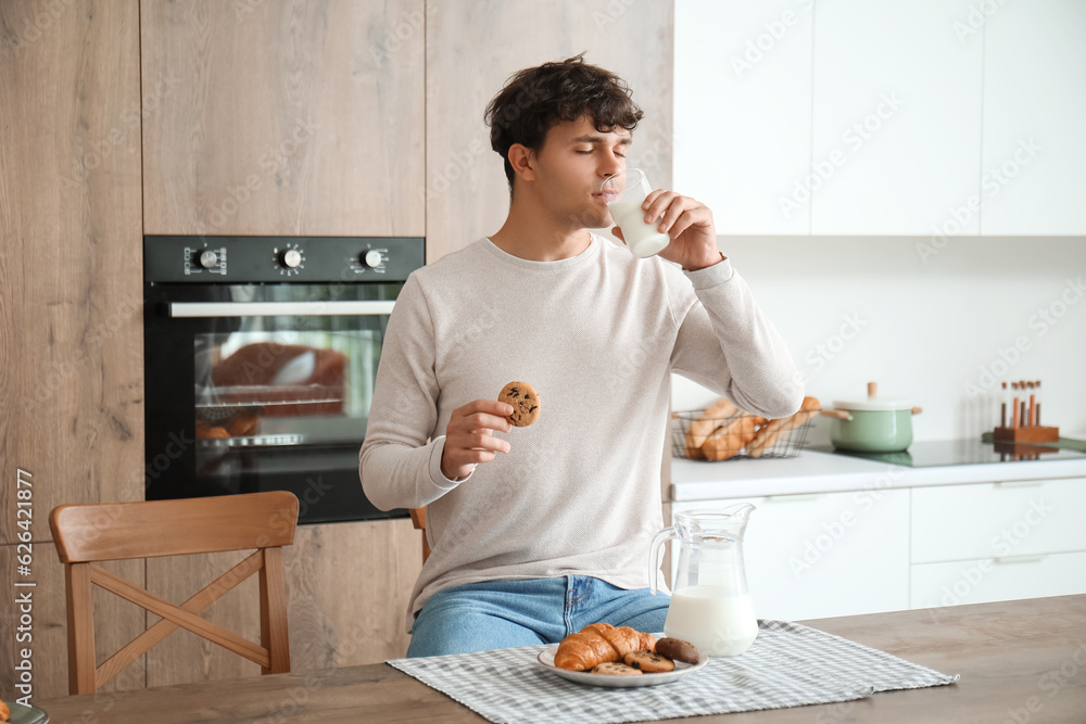 Young man drinking fresh milk and eating cookie in kitchen