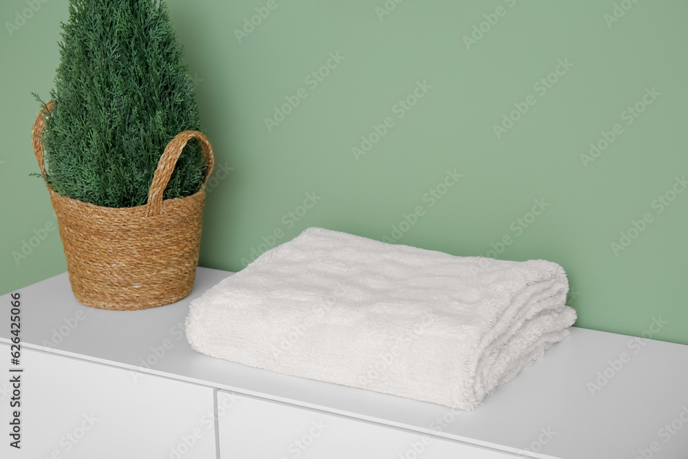 Folded blanket and wicker basket with plant on white cabinet near green wall