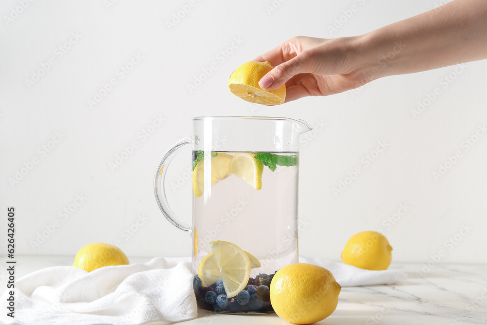 Woman squeezing lemon juice into jug of infused water with blueberries and blackberries on white tab