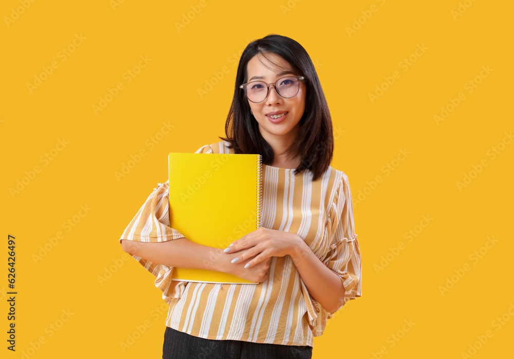 Female Asian teacher with notebook on yellow background