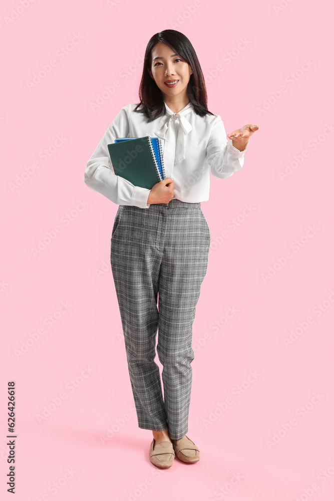 Female Asian teacher with notebooks on pink background