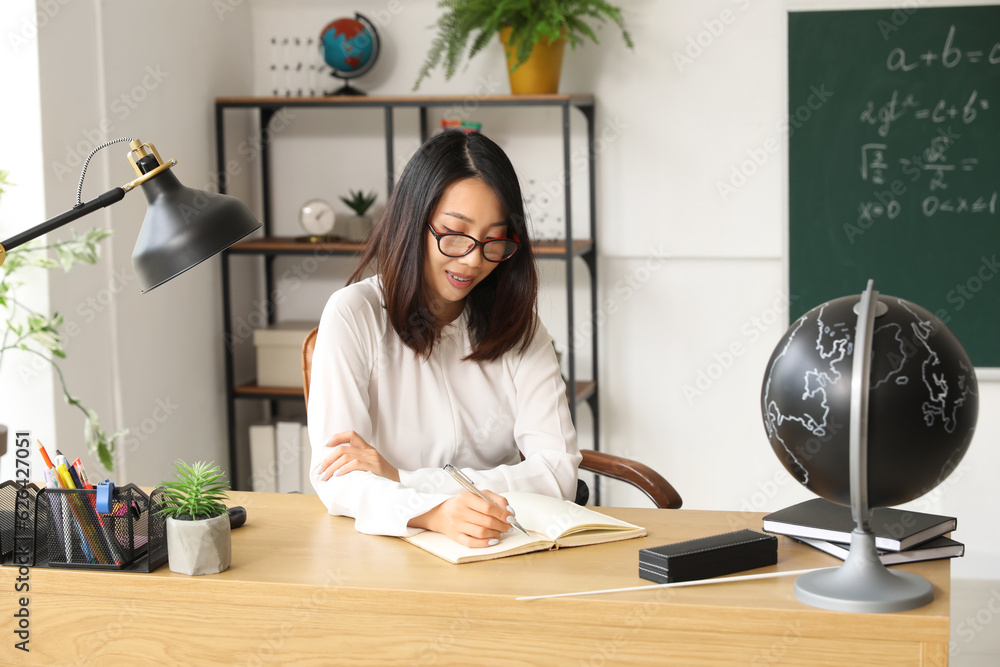 Female Asian teacher writing at table in classroom