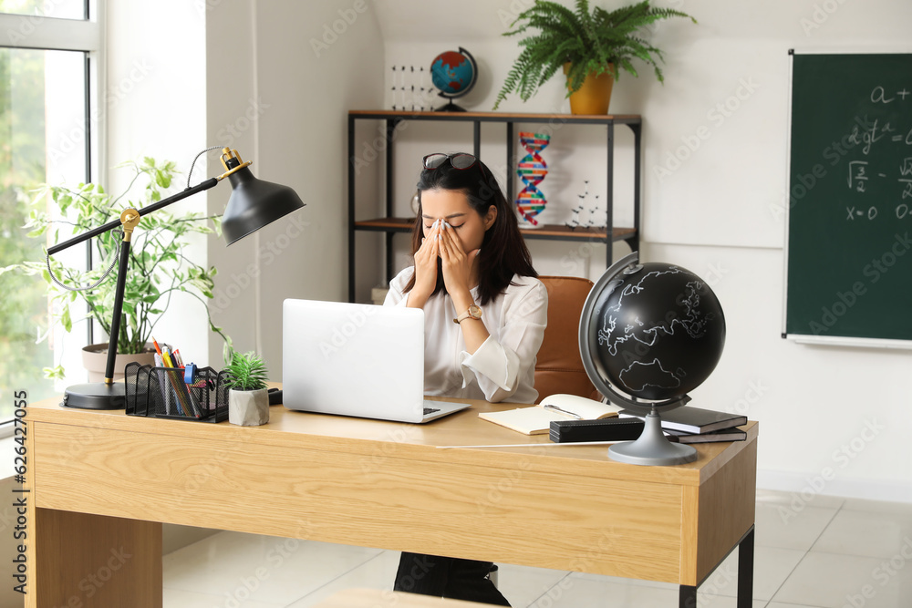 Tired Asian teacher working with laptop at table in classroom