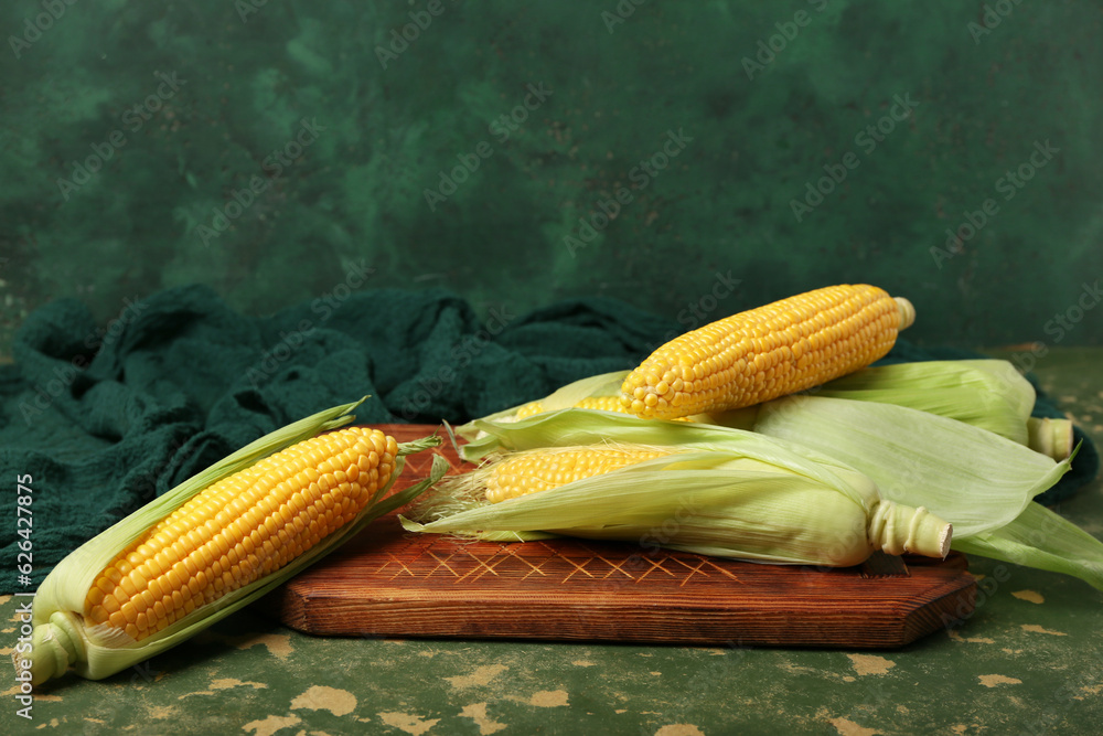 Wooden board with fresh corn cobs on green background