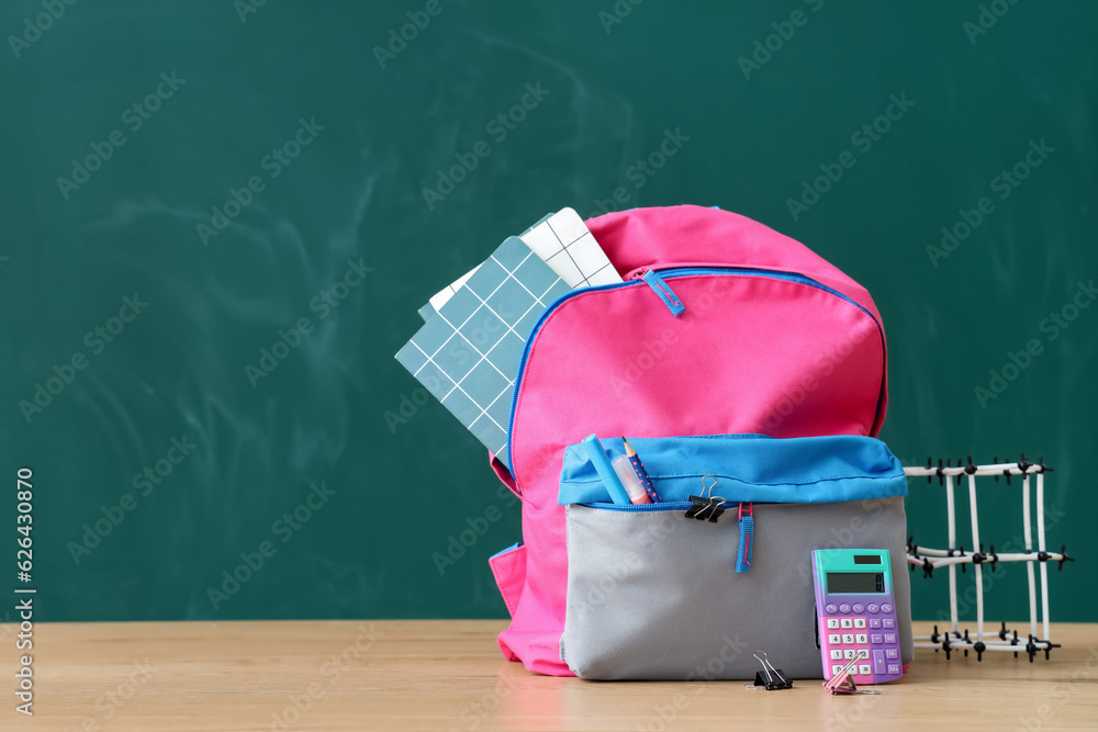 Backpack with school stationery and molecular model on table near green chalkboard