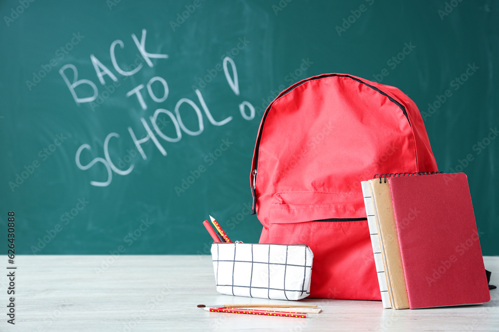 Backpack and supplies on table near green chalkboard with text BACK TO SCHOOL