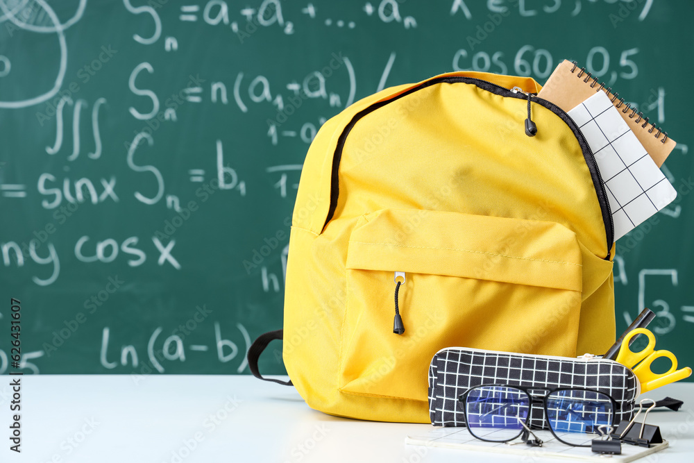 Yellow school backpack with eyeglasses and stationery on white table near green chalkboard