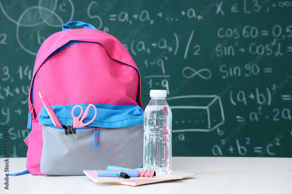 Colorful school backpack with bottle of water and stationery on white table near green chalkboard