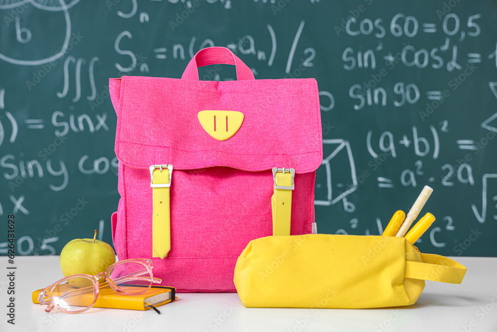 Pink school backpack with fresh apple and stationery on white table near green chalkboard