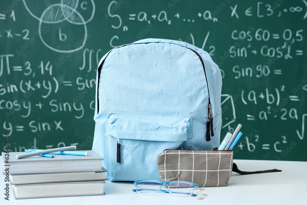 Blue school backpack with eyeglasses and stationery on white table near green chalkboard