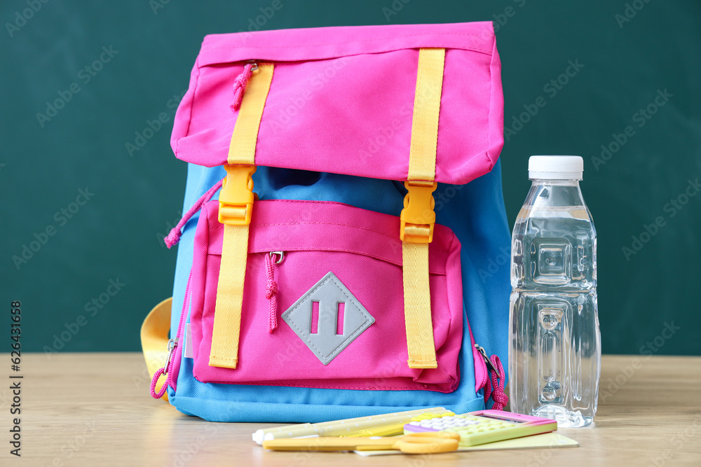 Colorful school backpack with stationery and bottle of water on wooden table near green chalkboard