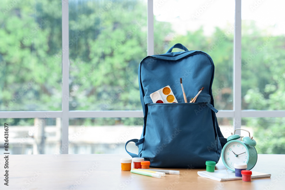 Blue school backpack with stationery and alarm clock on wooden table near window in classroom