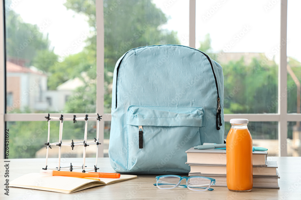 Blue school backpack with stationery, molecular model and bottle of juice on wooden table near windo