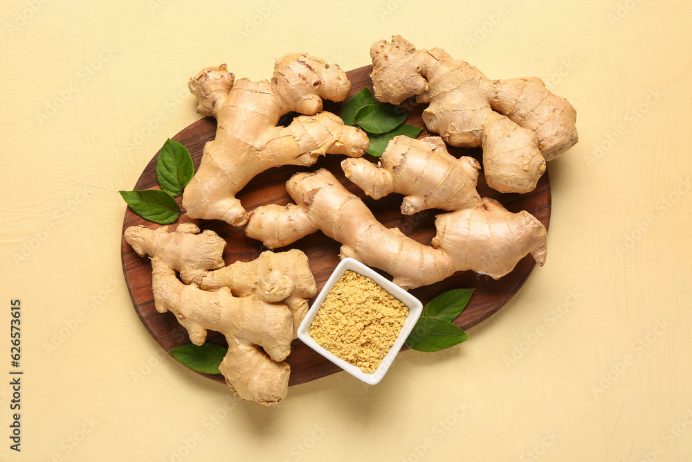 Wooden board with fresh ginger roots, leaves and bowl of dried powder on yellow background