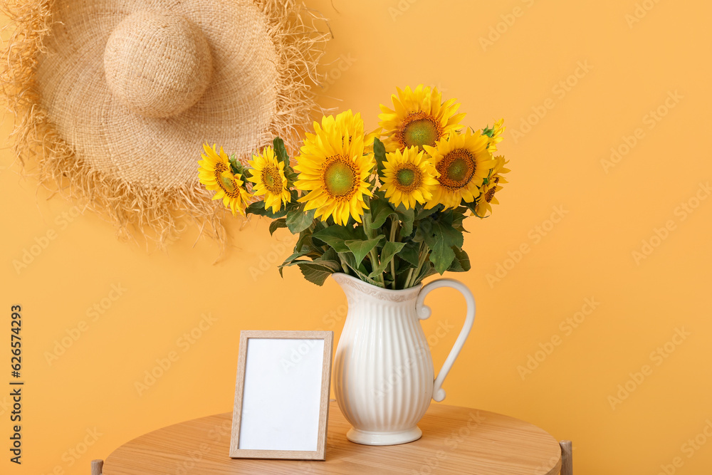 Vase with beautiful sunflowers, blank frame on table and wicker hat hanging on orange wall in room