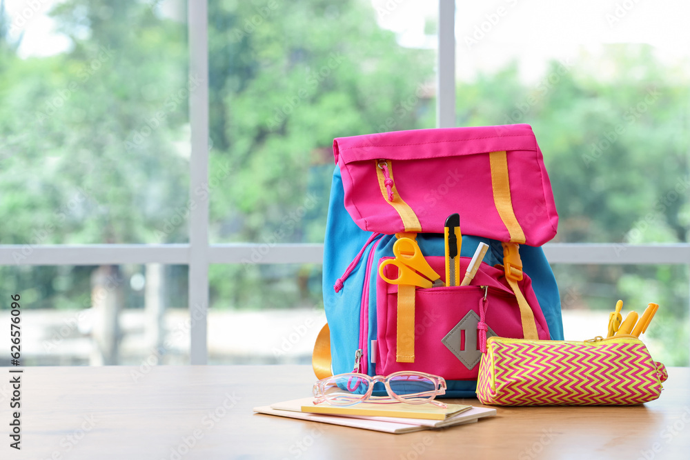 Colorful school backpack with stationery and eyeglasses on wooden table near window in classroom