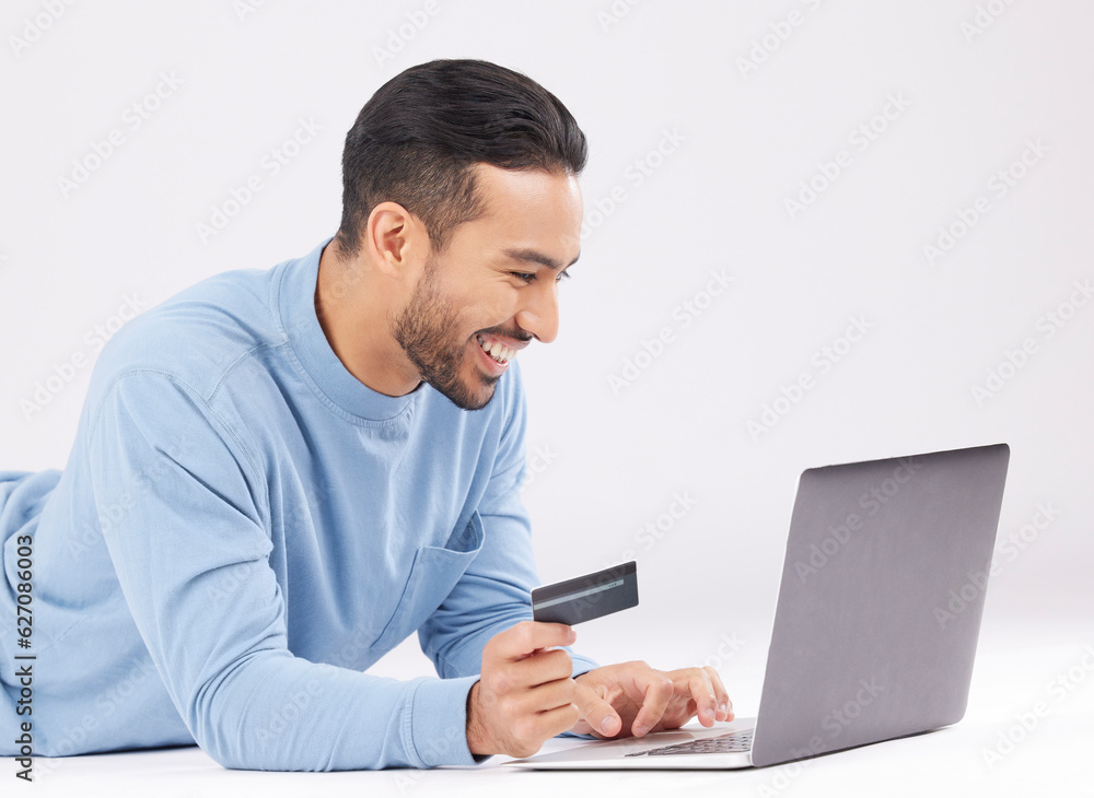 Laptop, credit card and happy asian man on studio floor for ecommerce, sale or cashback on grey back