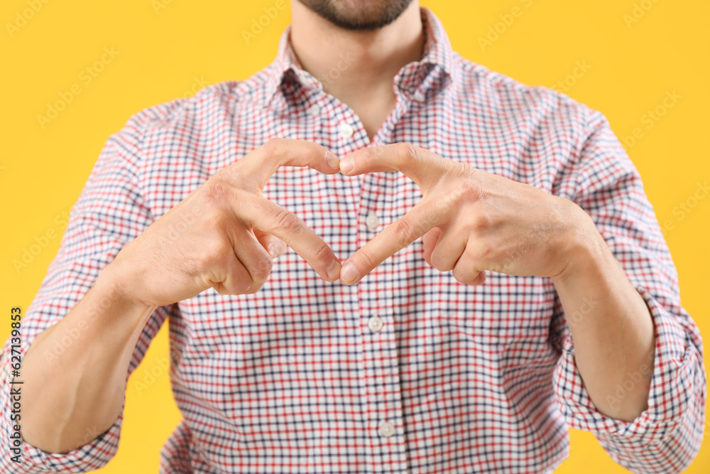 Handsome man making heart with his hands on yellow background, closeup