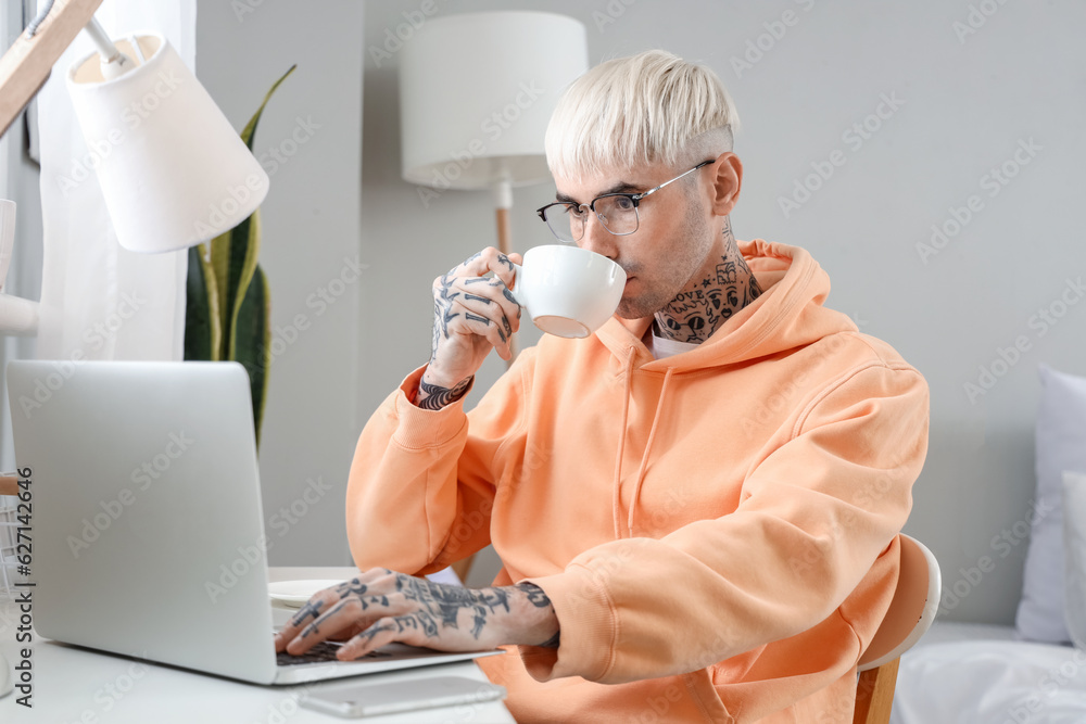 Young tattooed man in eyeglasses with laptop drinking coffee at home