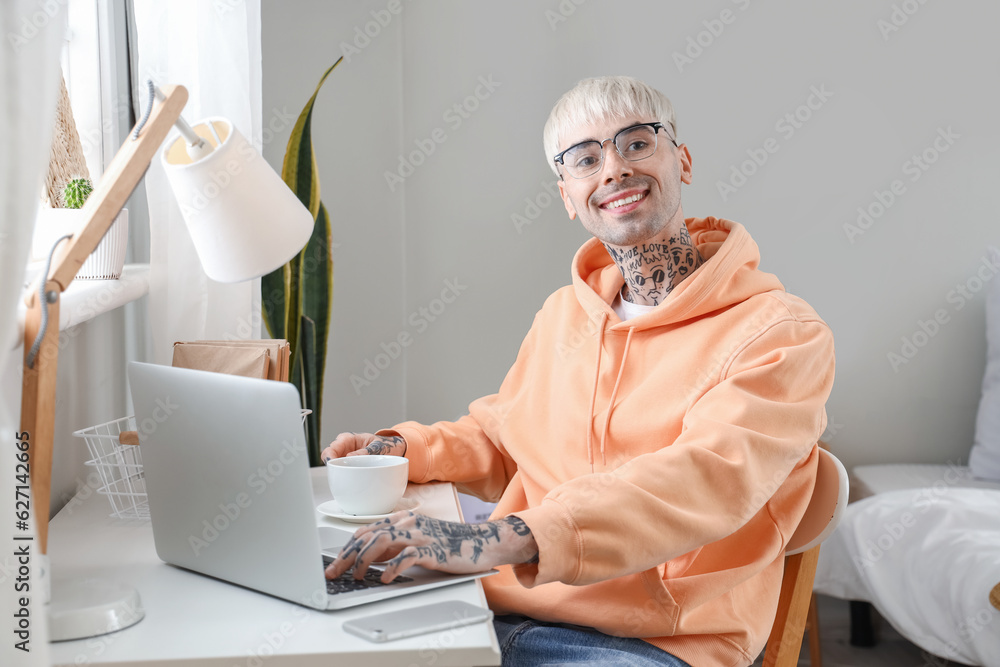 Young tattooed man in eyeglasses with laptop and cup of coffee at home