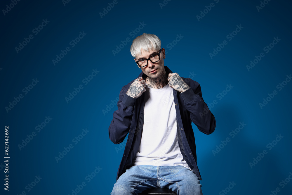 Young tattooed man in eyeglasses sitting on blue background