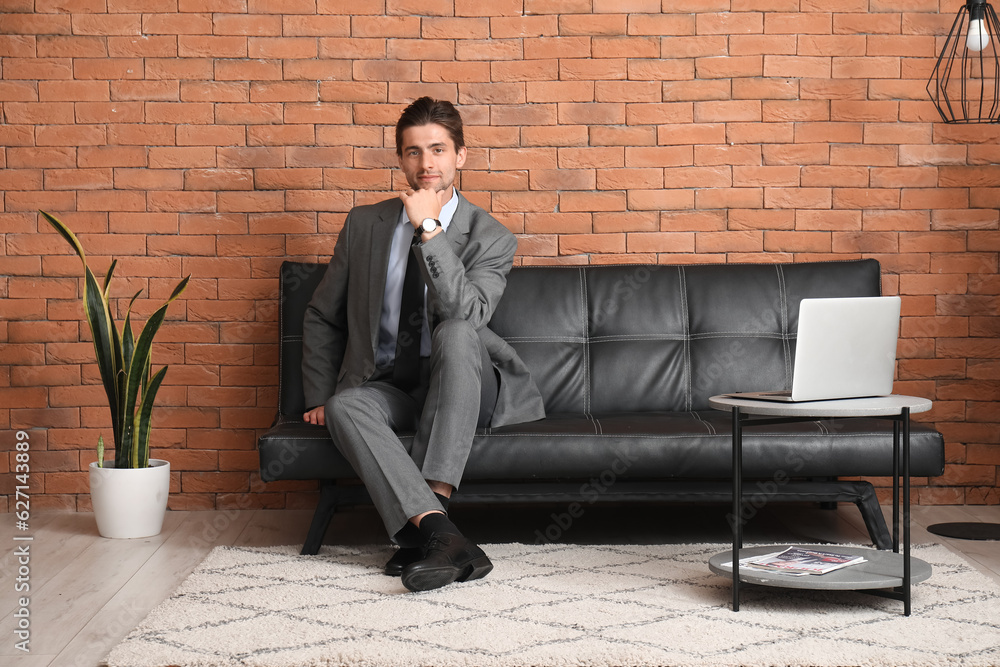 Stylish young man in suit with wristwatch and laptop sitting on leather sofa