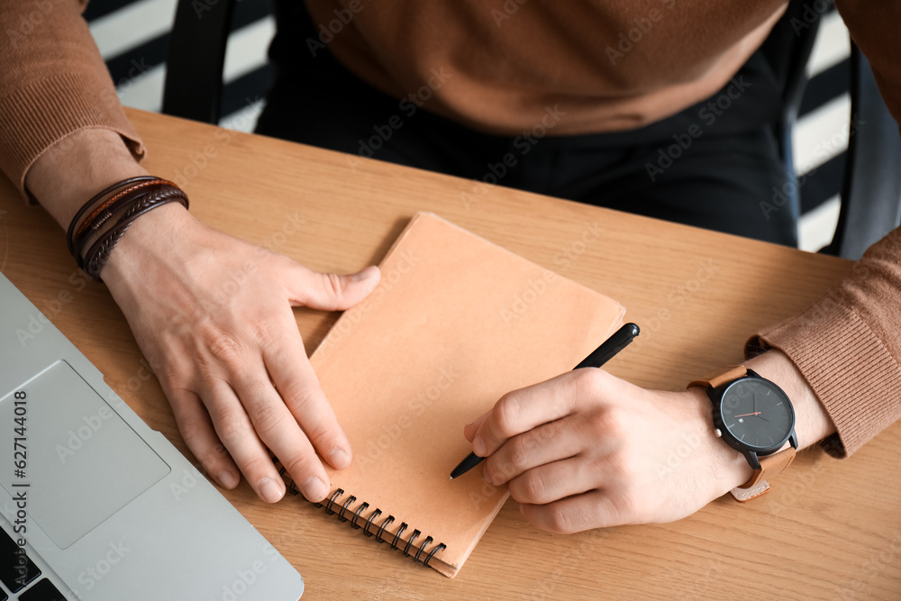 Stylish young man with wristwatch writing in notebook and sitting at office, closeup