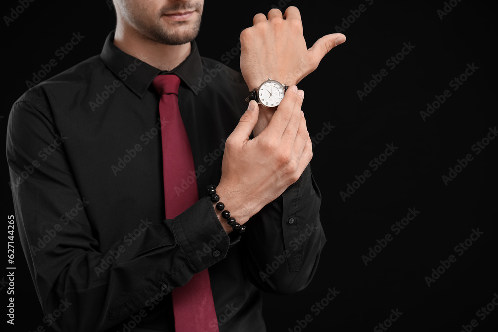 Elegant young man looking at wristwatch on black background, closeup