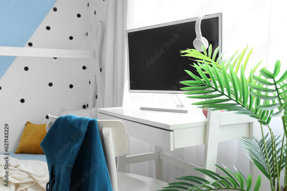 Desk with modern computer and houseplant in interior of childrens bedroom