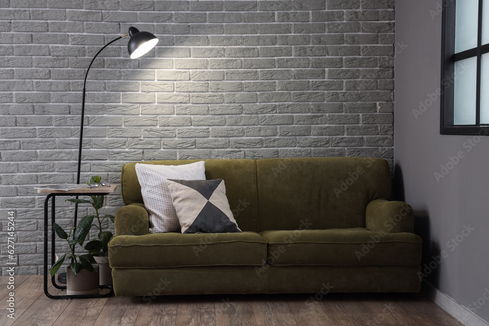 Glowing lamp and cozy green sofa near grey brick wall