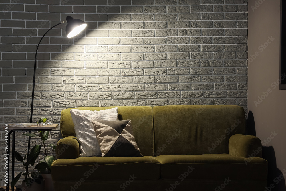 Glowing lamp and cozy green sofa near grey brick wall