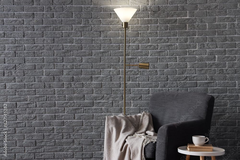 Glowing lamp and cozy armchair near grey brick wall