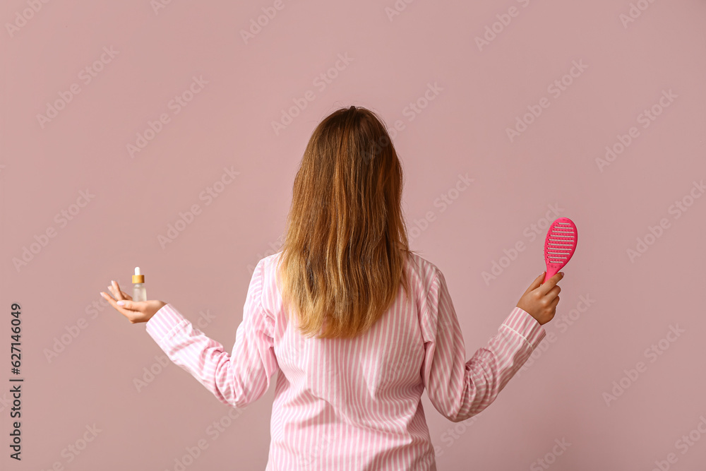 Young woman with cosmetic oil for hair treatment and brush on pink background, back view