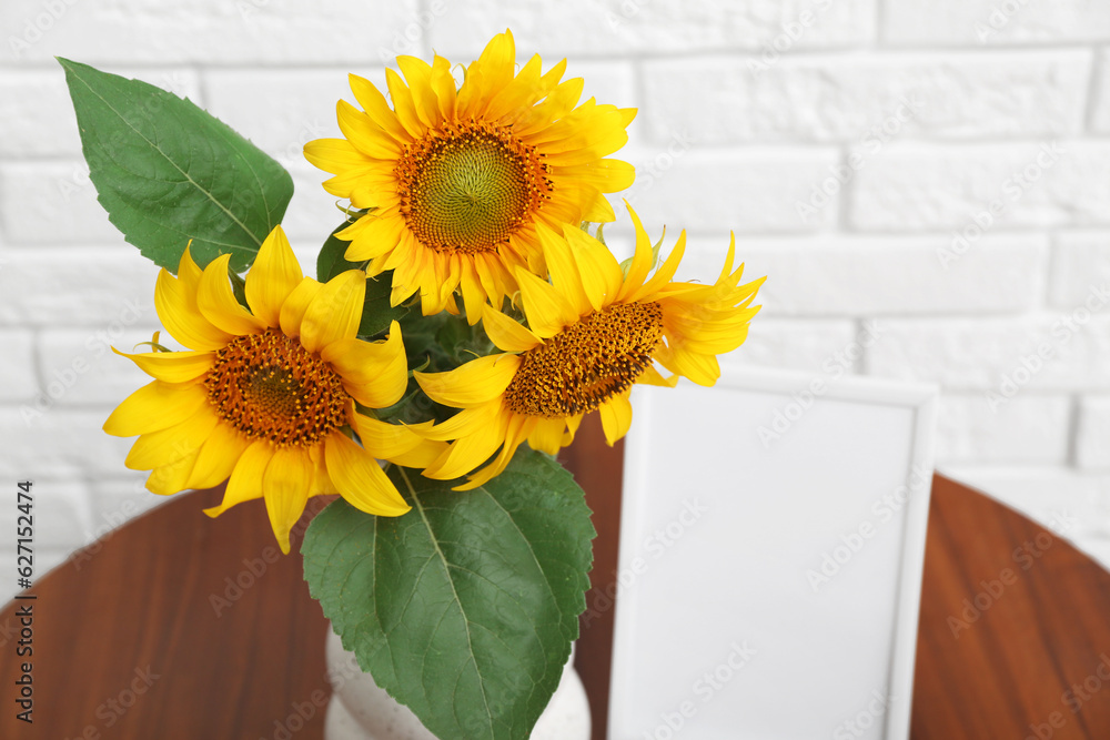 Vase with beautiful sunflowers and blank frame on table near light brick wall in room, closeup