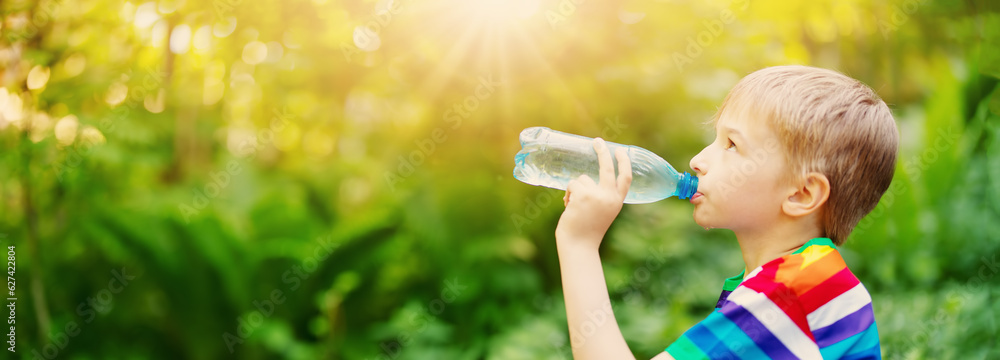 Boy standing in the natural park and drinking water from transparent bottle.