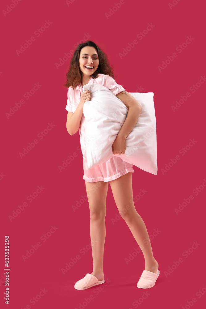 Young brunette woman in pajamas with pillow on pink background