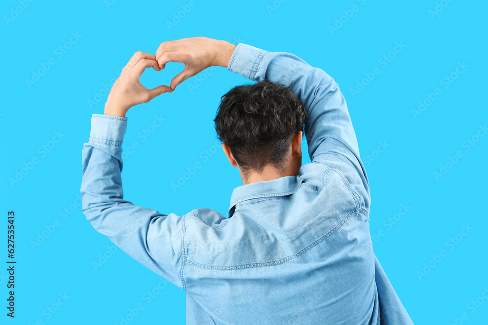 Handsome young man making heart with his hands on light blue background, back view